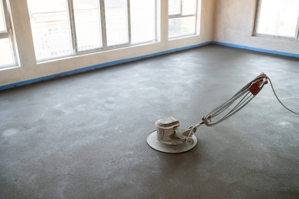 Floor grinding machine smoothing a concrete surface inside an empty room with large windows